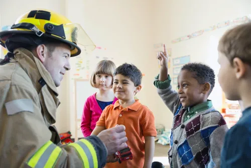 A firefighter in turnout gear teaches fire safety to elementary school students during a classroom visit